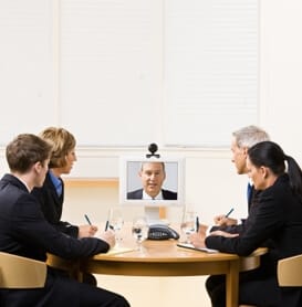 a group of employees at a conference table teleconferencing with a man in a business suit
