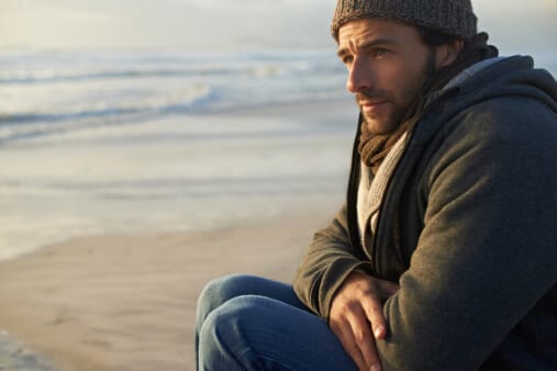 man with concerns about alcoholism bundled up on beach looking out to sea with arms folded