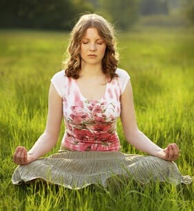young woman in dress seated in lotus position yoga posture in a green field