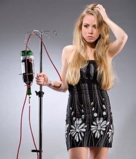 a confused looking young woman connected to an IV fluid bag that contains a bottle of wine