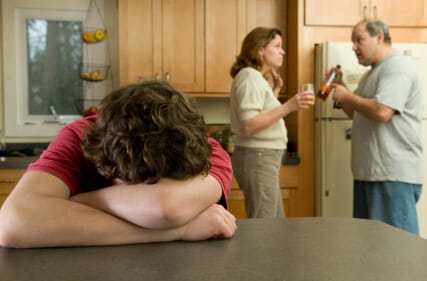 young man distraught at kitchen table as parents argue over bottle in background