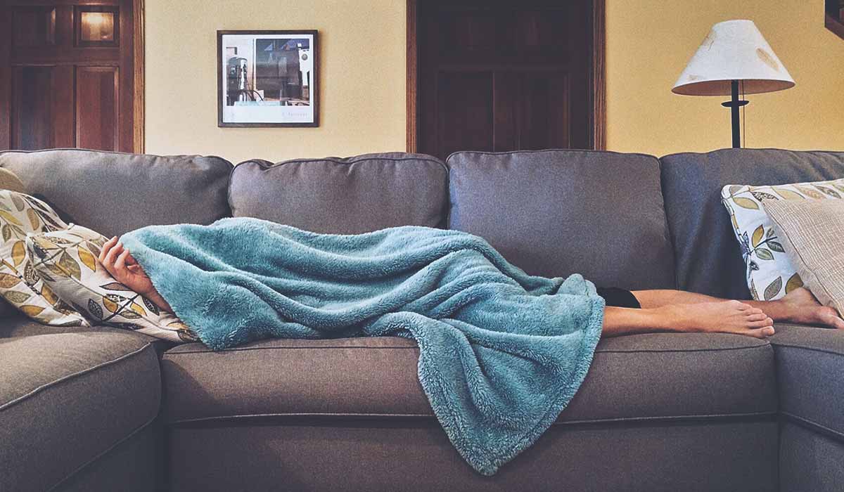 male flu sufferer on couch covered by blanket with only feet visible