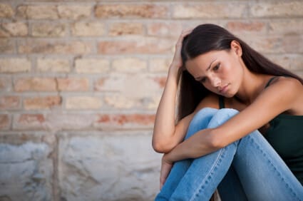 young woman with her arms around her knees looking down sadly next to brick wall
