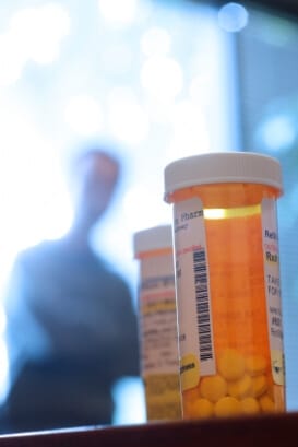 a closeup of prescription pill bottles in the foreground with the blurred image of a man with folded arms in the background demonstrating the dangers of prescription drug abuse