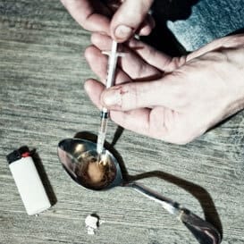 close up of hands drawing heroin into a syringe from a spoon featuring a disposable lighter and a small amount of the drug on the table next to it